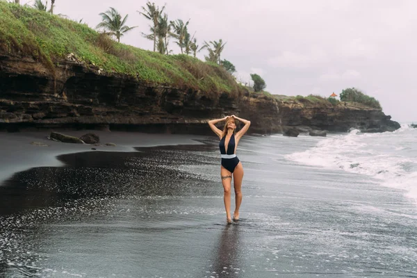 Κορίτσι με σέξι σώμα σε μαγιό περπατάει σε μαύρη αμμώδη παραλία. Σχέδιο ταξιδίου — Φωτογραφία Αρχείου