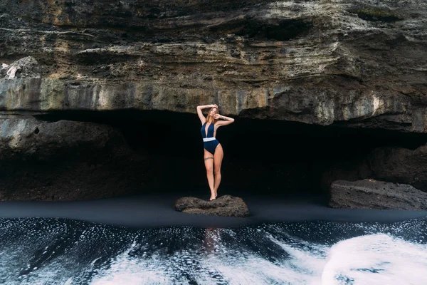 Sexy dívka se rozkládá v modrém plavkách blízko jeskyně na pláži s černým pískem, koncepce cestování — Stock fotografie