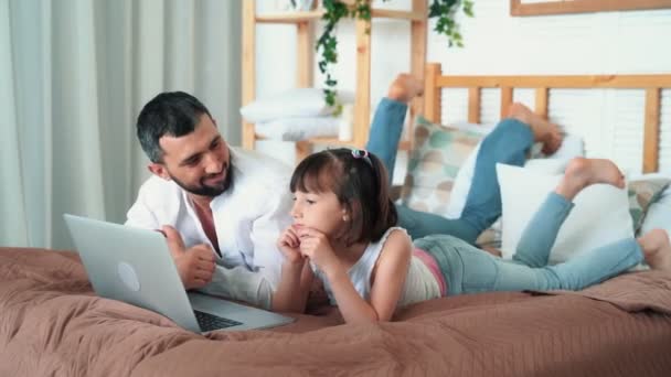 Papá y la niña linda ver dibujos animados en el ordenador portátil en casa se encuentra en la cama, cámara lenta — Vídeos de Stock