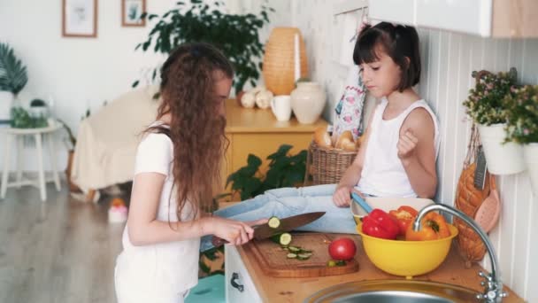 Маленька дівчинка ріже огірок на дошці для кухні, її сестра сидить на столі, повільний рух — стокове відео