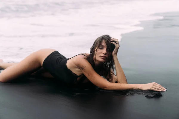 Sexy sensual girl with wet hair in swimwear lies on deserted black sandy beach — Stock Photo, Image