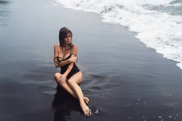 Sinnliches Mädchen im Badeanzug mit nassen Haaren sitzt am schwarzen Sandstrand in Meereswellen — Stockfoto