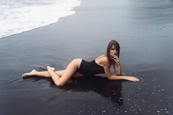 Sexy tanned girl with wet hair in swimwear lies on deserted black sandy beach — Stock Photo, Image