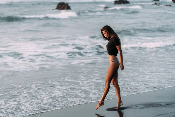 Chica con cuerpo sexy en camiseta negra y trajes de baño camina en la playa de arena negra —  Fotos de Stock