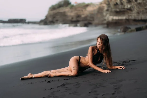 Sportig tjej med sexig kropp i svarta badkläder vilar på svart sandstrand — Stockfoto