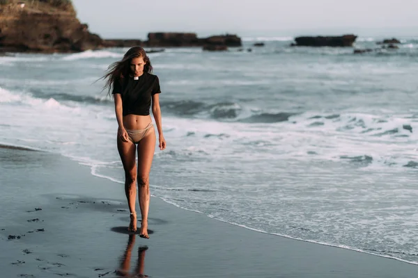 Chica con cuerpo sexy en camiseta negra y trajes de baño camina en la playa de arena negra —  Fotos de Stock
