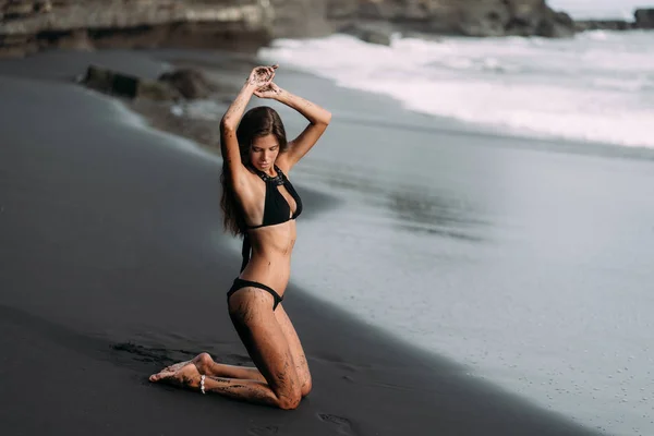 Young sexy girl in black swimwear resting on black volcanic sand beach — Stock Photo, Image