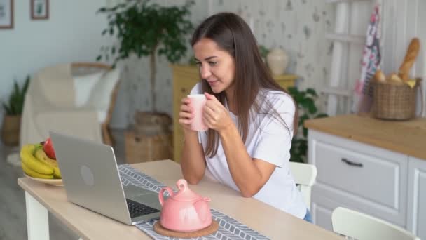 Jeune femme s'assoit à la cuisine, boit du thé et regarde un film sur ordinateur portable, ralenti — Video