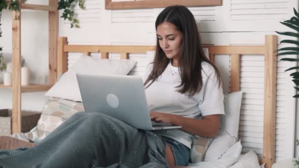 Bella ragazza si siede sul letto, utilizza il computer portatile per lavoro o chat online, rallentatore — Video Stock