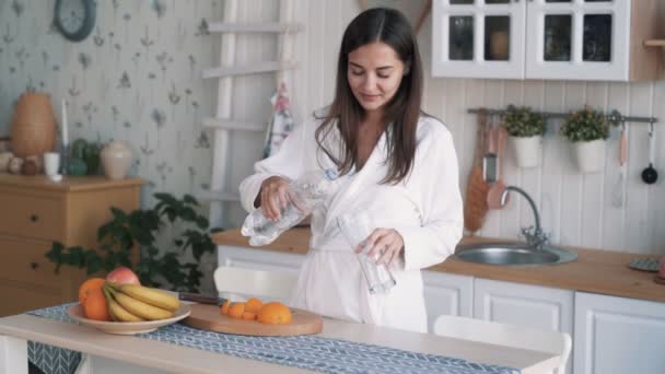 Menina em roupão derrama água limpa em vidro na cozinha, bebe, câmera lenta — Vídeo de Stock