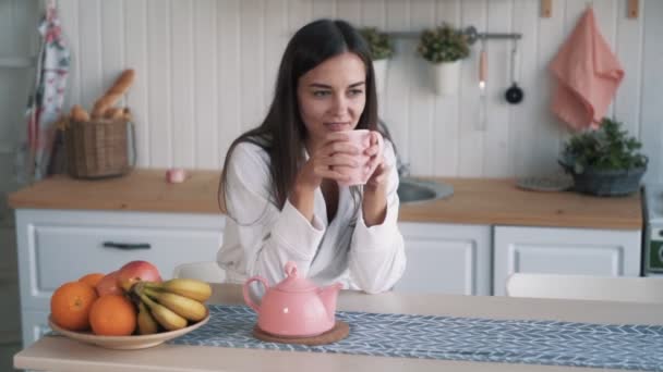 Hermosa chica sonriente bebe café en la cocina y mira hacia otro lado, cámara lenta — Vídeo de stock