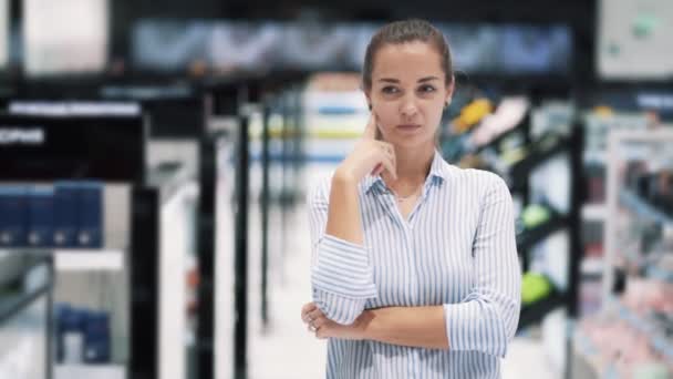 Chica en la tienda de cosméticos piensa qué producto comprar, hace cara divertida, cámara lenta — Vídeos de Stock