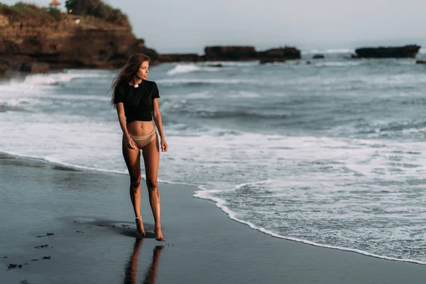 Chica en camiseta negra y traje de baño en la playa de arena negra en los rayos de la puesta del sol —  Fotos de Stock