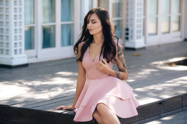 Красивая сексуальная девушка в розовом платье сидит на деревянных ступеньках уличного кафе — стоковое фото