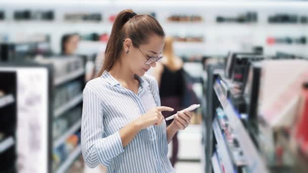Девушка в очках в косметическом магазине пользуется телефоном, смотрит на мобильный экран, замедленная съемка — стоковое видео