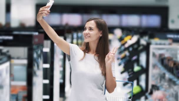 Girl makes selfie on phone in cosmetics store, shows peace gesture, slow motion — Stock Video