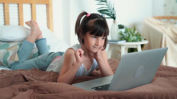 Niña acostada en la cama, utiliza el ordenador portátil, presiona los botones en el teclado, cámara lenta — Vídeos de Stock