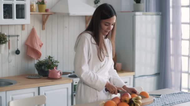 Niedliches Mädchen im weißen Bademantel schneidet Orange an Bord in der Küche, Zeitlupe — Stockvideo