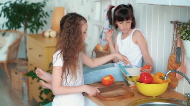 Meisje snijdt peper in de keuken, haar zus zit op tafel en eet Bun, Slow Motion — Stockvideo