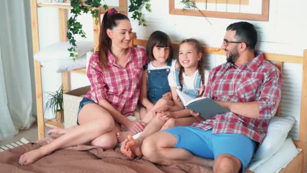 Eltern und zwei Töchter liegen im Bett und Papa liest ihnen Buch vor, Zeitlupe — Stockvideo