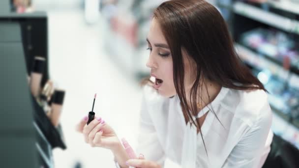 Menina bonita escolhe batom na loja de cosméticos, tenta em lábios, câmera lenta — Vídeo de Stock