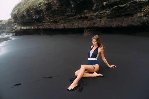 Ragazza sexy con grandi seni in costume da bagno blu rilassante in spiaggia con sabbia nera . — Foto Stock