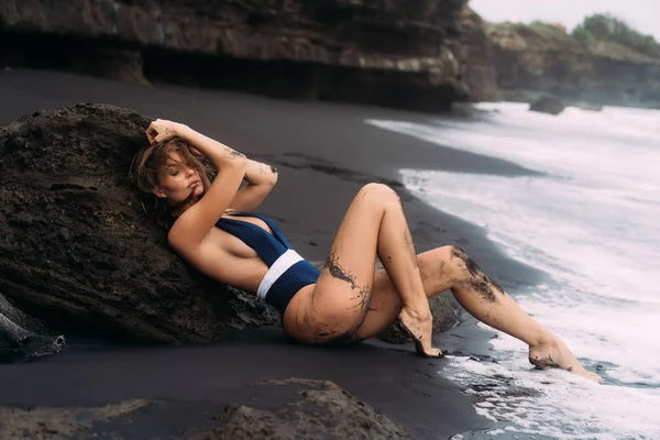 검은 모래 해변에서 편안한 블루 수영복에 큰 가슴을 가진 섹시 한 소녀. 로열티 프리 스톡 이미지