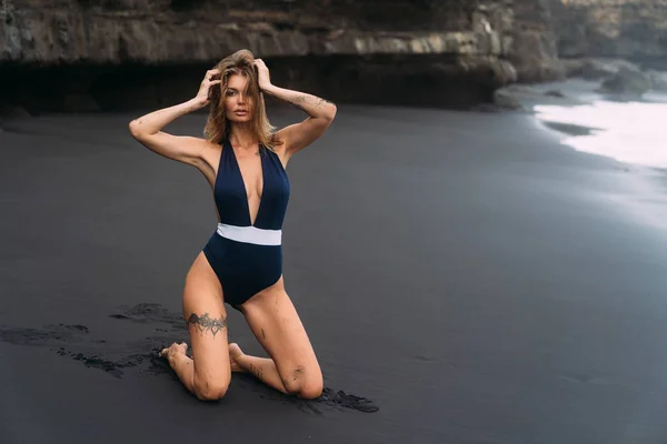 Menina sexy com seios grandes em roupa de banho azul relaxante na praia com areia preta . — Fotografia de Stock