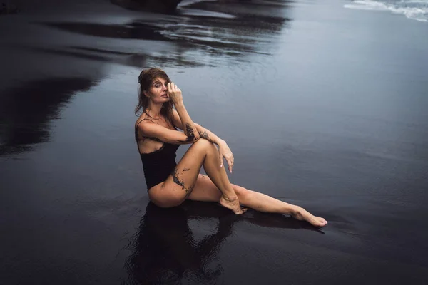 Sensuell sexig tjej i baddräkt med vått hår sitter på svart sandstrand — Stockfoto