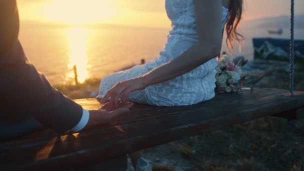Close up recém-casados de mãos dadas e balançar no balanço no fundo do pôr-do-sol dourado — Vídeo de Stock