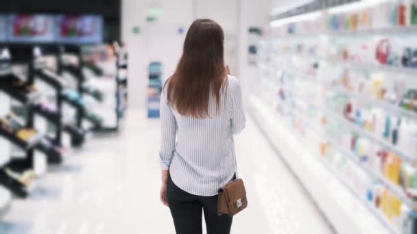 Задний вид, женщина идет среди полок в косметическом магазине, замедленная съемка — стоковое видео