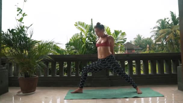 Mujer haciendo ejercicios de yoga en el balcón en la esterilla de yoga con vegetación en el fondo — Vídeos de Stock