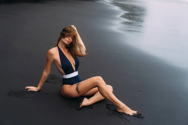 검은 모래 해변에서 편안한 블루 수영복에 큰 가슴을 가진 섹시 한 소녀. 스톡 이미지