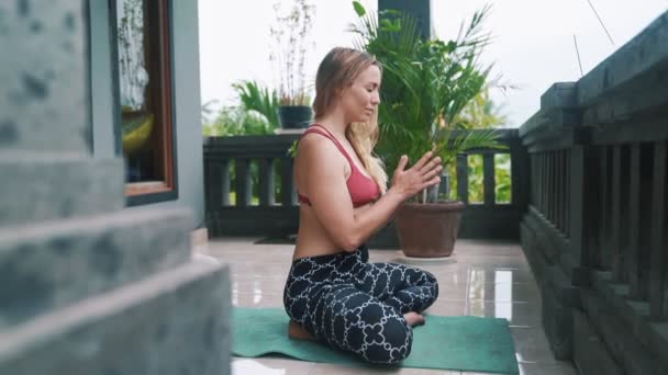 Young woman doing yoga exercises, meditates and prays on yoga mat on veranda — Stock Video