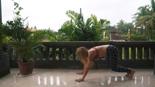 Молодая женщина практикует йогу позировать на балконе с пальмой на заднем плане — стоковое видео