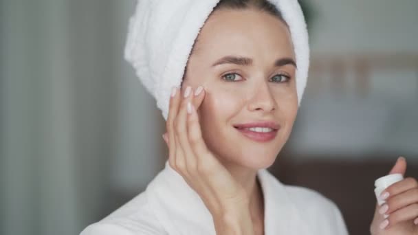 Kvinna med handduk på huvudet applicerar fuktighetskräm grädde på hennes ansikte, närbild — Stockvideo
