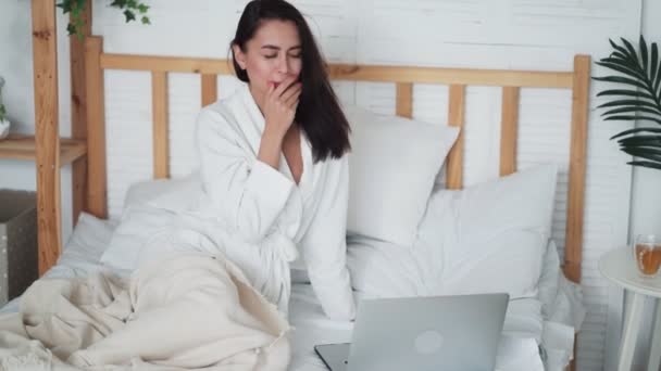 Mujer joven en albornoz se sienta en la cama, utiliza el ordenador portátil para el trabajo en casa y bosteza — Vídeos de Stock