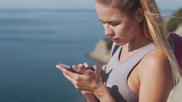 Porträtt av idrottskvinna i sportkläder skickar meddelande på telefon, havet på bakgrunden — Stockvideo