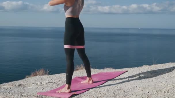 Schlanke Frau duckt sich auf Matte mit Gummiband im Freien in der Nähe des Meeres, Zeitlupe — Stockvideo