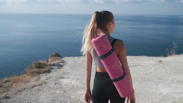 Rückansicht, Mädchen in Sportbekleidung mit Gymnastikmatte geht auf den Gipfel des Berges — Stockvideo