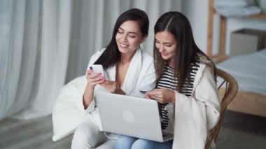 Yatak odasında sandalyede oturan iki kadın dizüstü bilgisayar, telefon, kart ve internetten alışveriş yapıyor.