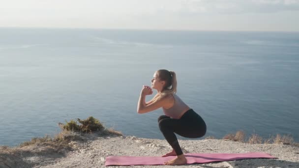 Vrouw maakt hurken op mat buiten, traint haar lichaam, met uitzicht op zee, slow motion — Stockvideo