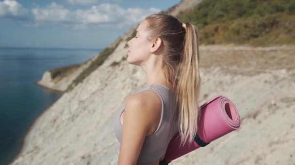 Frau hält Yogamatte, blickt von Klippe auf Meer, atmet tief, Zeitlupe — Stockvideo