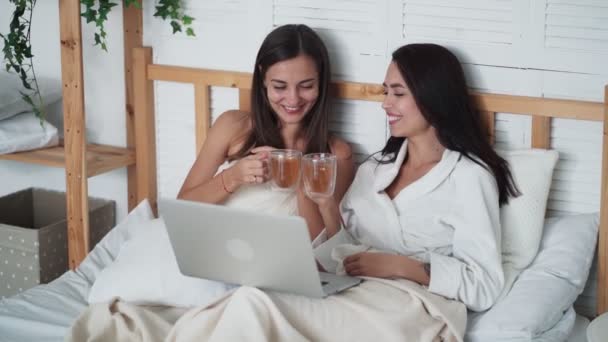 Due amiche si siedono a letto, bevono tè, guardano film divertenti sul computer portatile, ridono — Video Stock