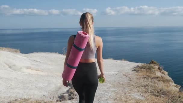 Backside view, kvinna i sportkläder med gymnastikmatta går till kanten av klippan — Stockvideo