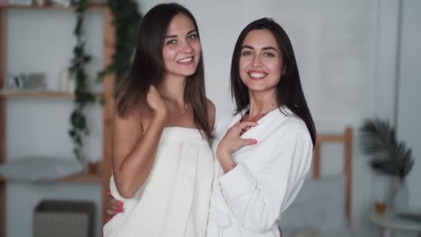 Retrato de dos hermosas mujeres en albornoces mirar a la cámara, sonrisa, en el dormitorio — Vídeos de Stock