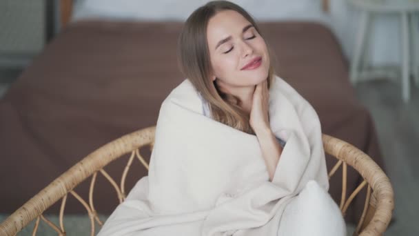 Beautiful young woman sits in chair wrapped in plaid, smiles and looks at camera — Stock Video