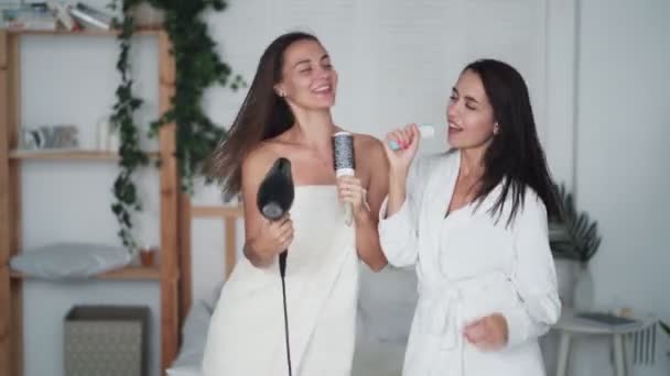 Två flickvänner i vit badrock dans, sjunga och torrt hår med hårtork, skratta — Stockvideo