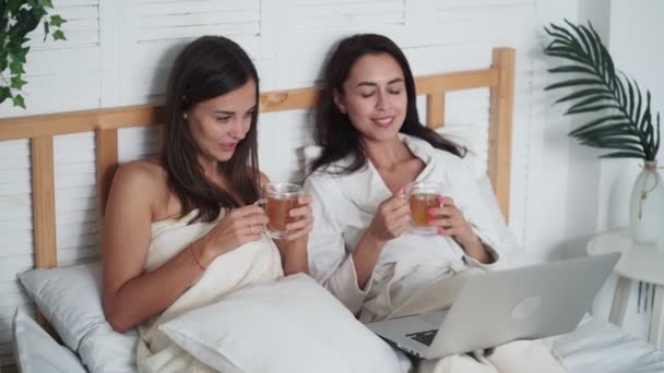 Två kvinnor vänner sitta i sängen, dricka te, titta på film på laptop, prata — Stockvideo