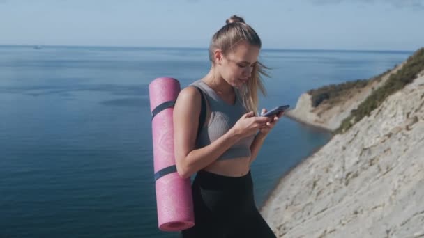 Спортсменка в спортивной одежде с ковриком отправляет сообщение по телефону, море на заднем плане — стоковое видео